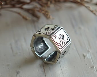 Puede incluir: Un anillo de plata con un diseño de cuatro cartas de juego, cada una con un palo diferente: diamantes, tréboles, corazones y picas. El anillo está hecho de metal y tiene una superficie texturizada.