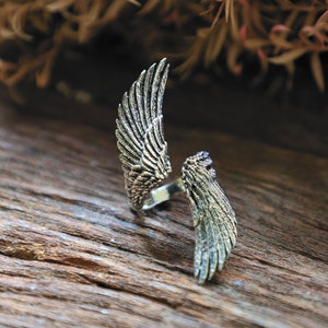 May include: A silver-toned ring with an open design, featuring detailed wing motifs. The ring's wings are spread open, showcasing intricate feather detailing. The ring is set against a rustic wooden surface, highlighting its metallic sheen.