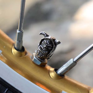 Puede incluir: Una tapa de válvula plateada con un diseño de parca está montada en una rueda de motocicleta dorada. La rueda tiene radios plateados y una franja blanca. La tapa presenta una cara y una capucha detalladas.