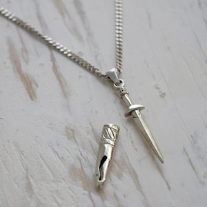 Puede incluir: Un collar de cadena de plata con un colgante de daga de plata y una funda de plata a juego. El colgante está detallado con una punta puntiaguda y una guarda cruzada.
