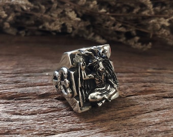 Könnte beinhalten: Silberfarbener Ring mit detaillierter Darstellung einer gehörnten Figur mit Flügeln, die einen Stab hält. Der Ring hat ein rechteckiges Design mit aufwendigen Seitenschnitzereien, vor rustikalem Holzgrund.