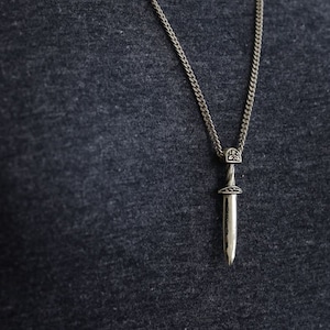 Puede incluir: Un collar de cadena de plata con un colgante de espada. La espada tiene un diseño detallado y una punta puntiaguda.