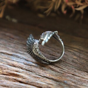 May include: A silver-colored ring with an eagle wing design. The ring features detailed feather textures and is set against a wooden background. The wing wraps around the finger, creating a unique and stylish accessory.