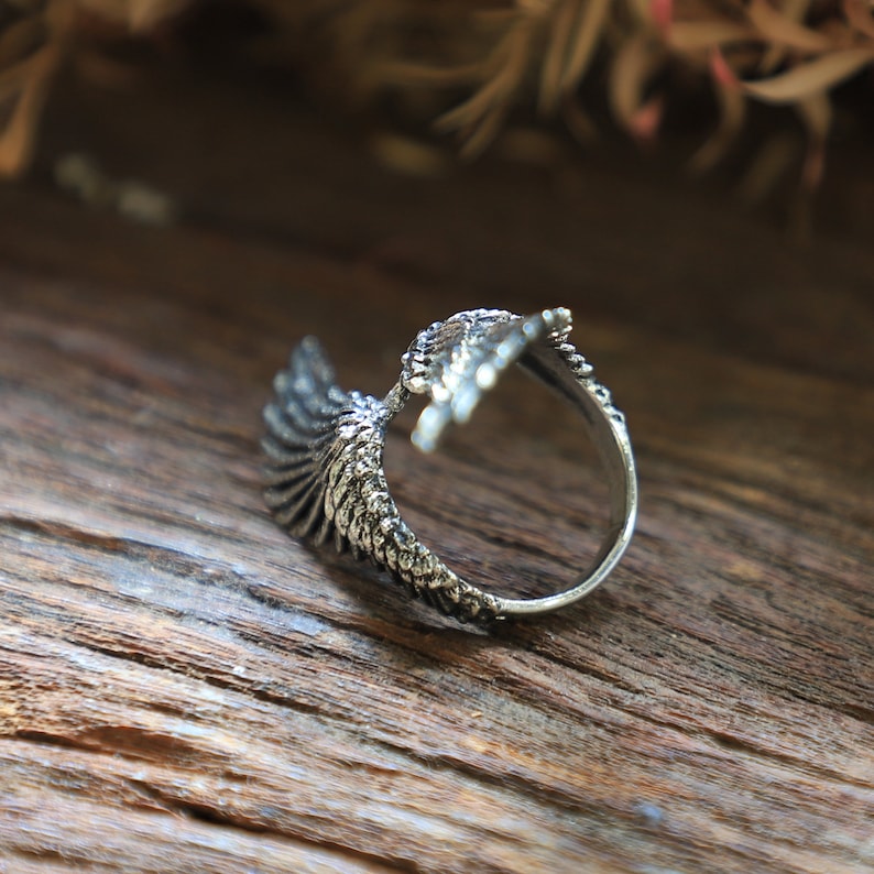 May include: A silver-colored ring with an eagle wing design. The ring features detailed feather textures and is set against a wooden background. The wing wraps around the finger, creating a unique and stylish accessory.