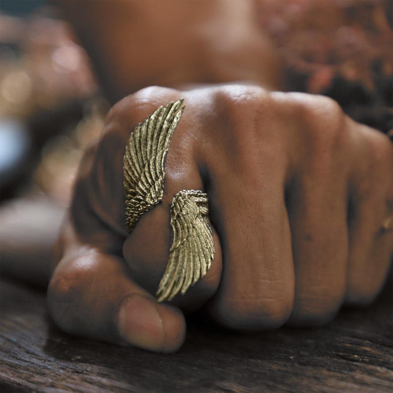 May include: A gold-colored ring shaped like two wings, one on each side of the finger. The ring is detailed with feather-like textures, creating a realistic appearance. The ring is worn on a hand with a blurred background.
