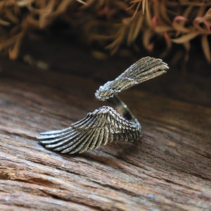 May include: A silver-colored ring with an open design, featuring detailed wing motifs. The ring's wings are spread out, creating a unique and artistic look. The jewelry is set against a wooden surface.