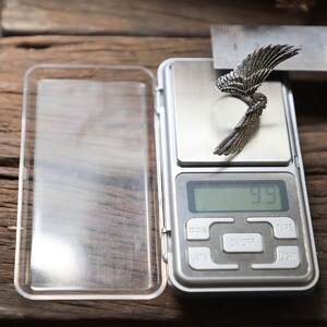 May include: A silver-colored wing-shaped ring is displayed on a digital scale, with a measurement of 9.9. The ring is positioned next to a ruler, suggesting a measurement is being taken. The scale is on a wooden surface.