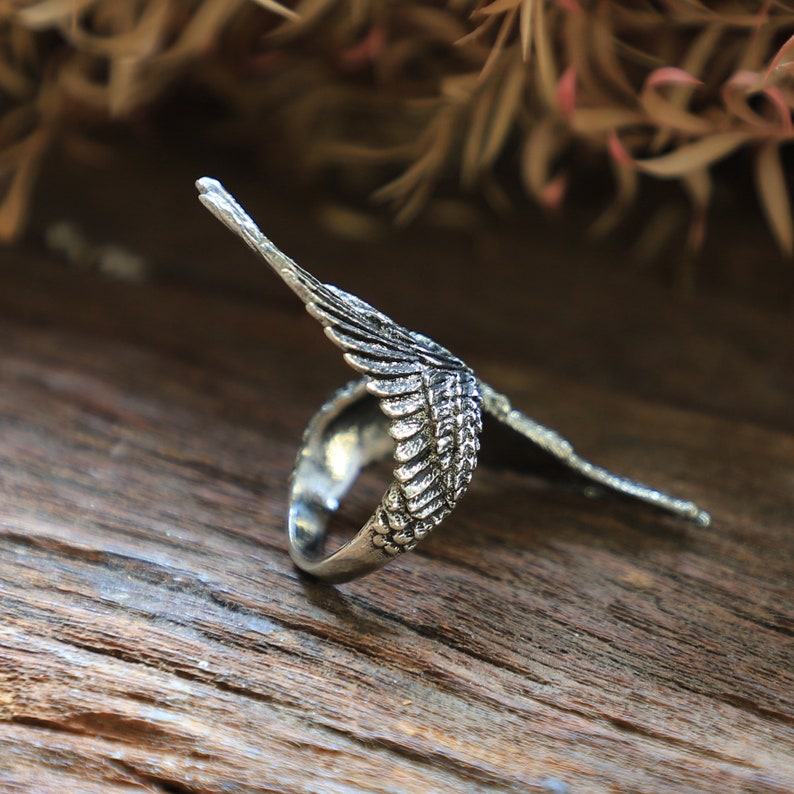 May include: A silver-colored ring with a detailed wing design. The ring features a textured surface, with the wing extending upwards. The ring is set against a wooden background, creating a contrast between the metal and the natural wood.