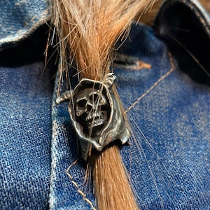 Peut inclure: Une bague argentée ornée d'un crâne avec une cape à capuche. La bague est mise en valeur sur un fond de tissu en jean et de cheveux châtain clair.