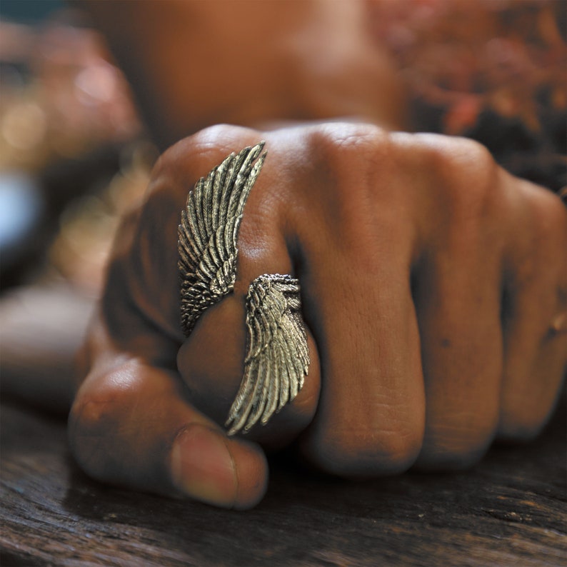 May include: A silver-colored ring with an open design, featuring detailed wing motifs on each side. The ring is worn on a hand, showcasing the intricate feather detailing and metallic sheen. The ring's design suggests a theme of freedom or flight.