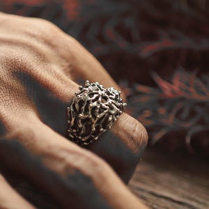 May include: A silver ring featuring an elaborate, organic pattern reminiscent of coral or a network of branches. The ring is worn on a finger, highlighting its detailed texture and unique form. The jewellery piece has a textured, openwork design.