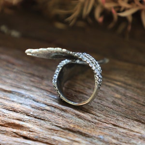 May include: A silver-toned ring with a textured band and a leaf-like design extending from the top. The ring is set against a wooden surface, showcasing its intricate details and craftsmanship.