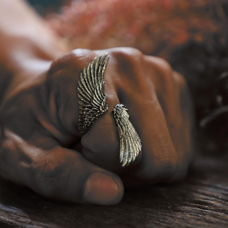 May include: A bronze-colored ring in the shape of angel wings. The ring features detailed feather textures and wraps around the finger. The wings are slightly open, creating a unique and artistic design. The ring is a statement piece.