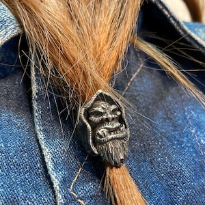 Peut inclure: Une bague argentée représentant un visage stylisé détaillé avec une barbe et une capuche. La bague est mise en valeur sur un fond de cheveux châtain clair et une veste en jean bleue.