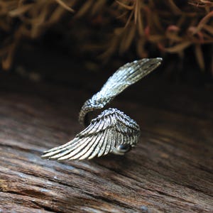 May include: A silver-toned ring shaped like angel wings. The ring features detailed feather textures and wraps around the finger. The wings are slightly open, creating a dynamic and elegant design. The ring is set against a rustic wooden surface.
