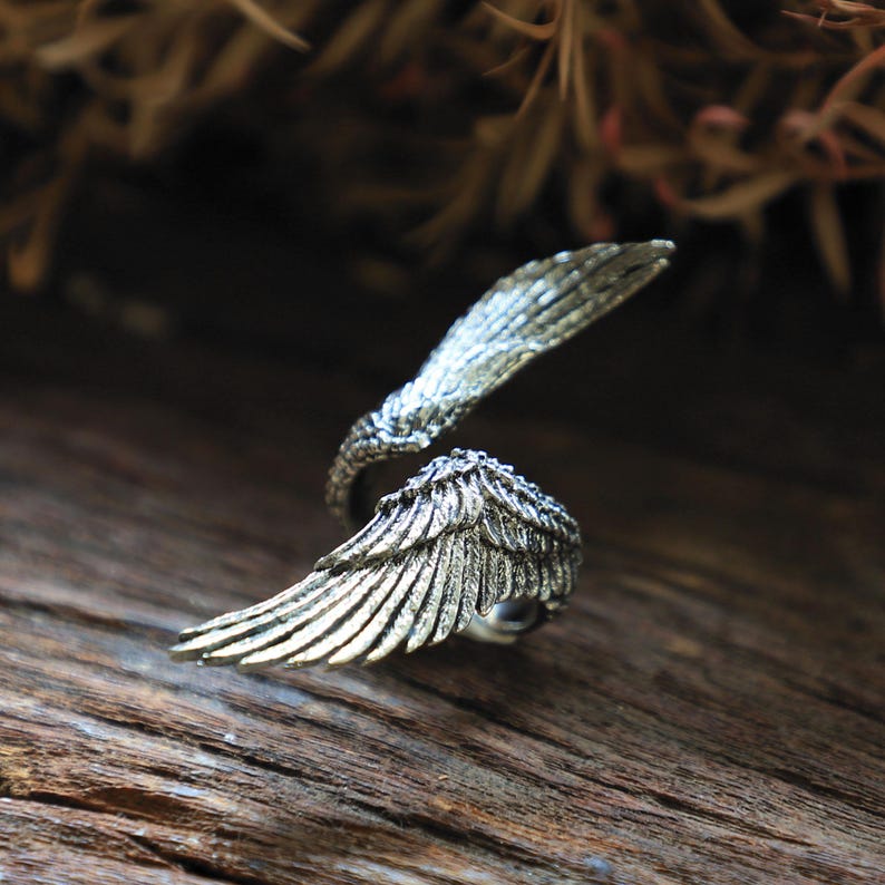 May include: A silver-toned ring shaped like angel wings. The ring features detailed feather textures and wraps around the finger. The wings are slightly open, creating a dynamic and elegant design. The ring is set against a rustic wooden surface.