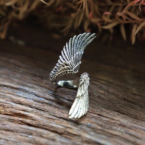 May include: A silver-colored ring with a wing design. The ring features two detailed wing sections, one forming the main part of the ring and the other extending outwards. The wings have intricate feather detailing, creating a textured appearance.