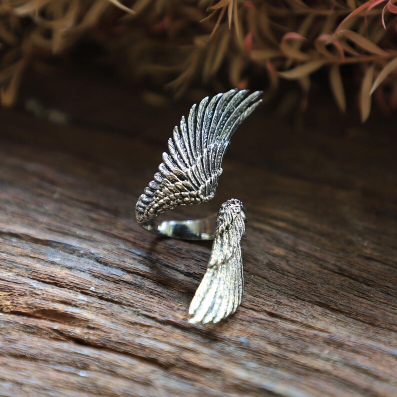 May include: A silver-colored ring with a wing design. The ring features two detailed wing sections, one forming the main part of the ring and the other extending outwards. The wings have intricate feather detailing, creating a textured appearance.