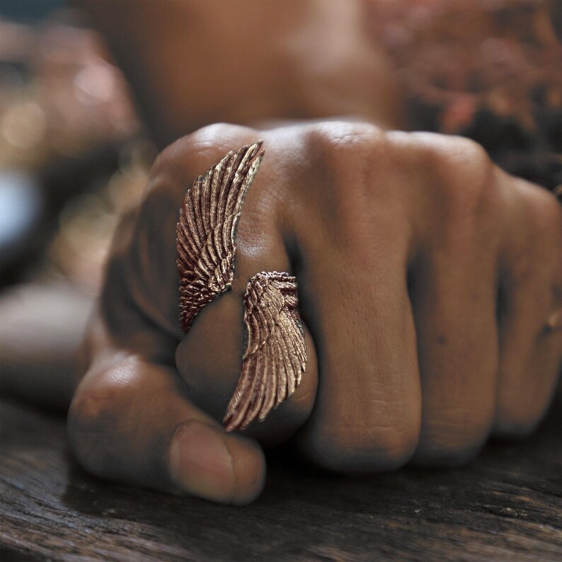 May include: A bronze-colored ring with a wing design. The ring wraps around the finger, with one wing on top and another on the side. The ring is detailed with feather-like textures.
