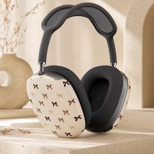 May include: Over-ear headphones featuring a cream-coloured exterior shell decorated with a repeating pattern of small brown bows. The device includes a dark grey mesh headband, dark grey ear cushions, and metallic silver adjustment arms.