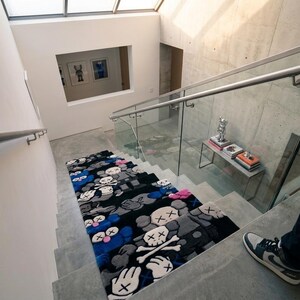 May include: A rectangular rug featuring a repeating pattern of cartoon-style characters with X-shaped eyes and skull motifs in shades of black, gray, blue, and pink, laid out on a concrete staircase with a glass railing and metal handrail.