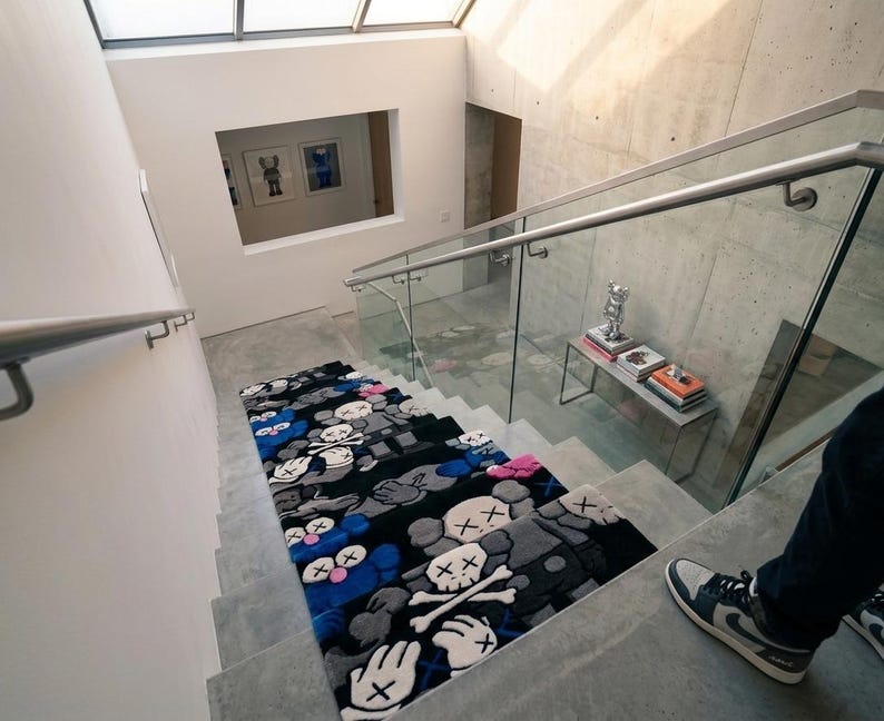 May include: A rectangular rug featuring a repeating pattern of cartoon-style characters with X-shaped eyes and skull motifs in shades of black, gray, blue, and pink, laid out on a concrete staircase with a glass railing and metal handrail.