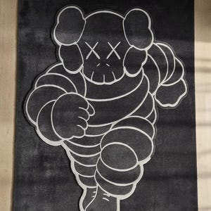 Puede incluir: Una alfombra en blanco y negro con una figura de dibujos animados estilizada con ojos tachados. La alfombra tiene un fondo gris oscuro con un contorno blanco del personaje. El dise&ntilde;o es de estilo gr&aacute;fico moderno.