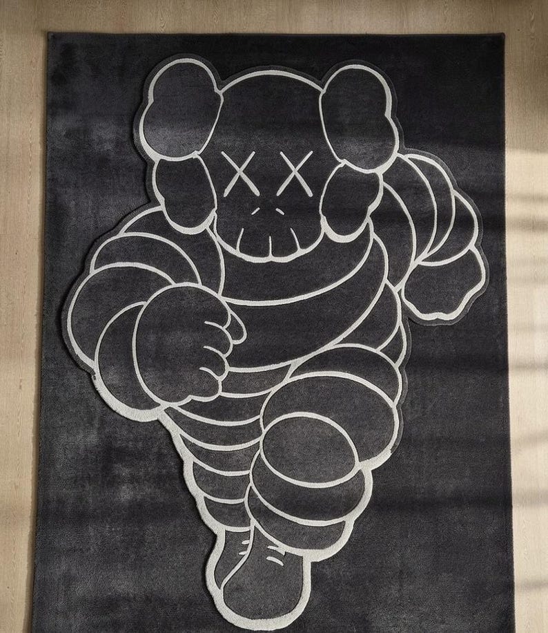 Puede incluir: Una alfombra en blanco y negro con una figura de dibujos animados estilizada con ojos tachados. La alfombra tiene un fondo gris oscuro con un contorno blanco del personaje. El dise&ntilde;o es de estilo gr&aacute;fico moderno.