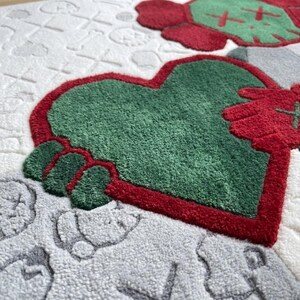 May include: A tufted rug featuring a large green heart held by red hands with cross-stitch eyes. The background is white with embossed, subtle patterns of skulls and cross-stitch motifs. A red and green skull figure is visible in the upper corner.