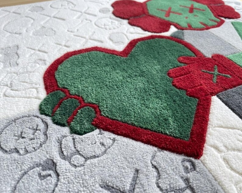 May include: A tufted rug featuring a large green heart held by red hands with cross-stitch eyes. The background is white with embossed, subtle patterns of skulls and cross-stitch motifs. A red and green skull figure is visible in the upper corner.