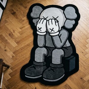 以下が含まれることがあります： グレーの体、白い手袋、大きな耳を持つスタイライズされた漫画のキャラクターが描かれたカスタム形状のラグ。キャラクターは顔を両手で覆って座っており、グレー、黒、白の色合いで太い輪郭線で表現されています。