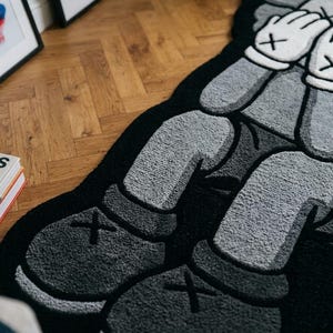 以下が含まれることがあります： グレーの肌、白い手袋、ダークグレーの靴を履いたデフォルメされた漫画のキャラクターが描かれたタフテッドラグ。キャラクターは座った姿勢で両手で顔を覆っており、黒い背景に黒い輪郭線で縁取られている。