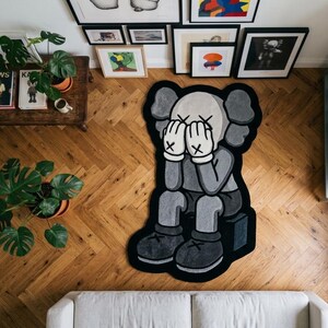 以下が含まれることがあります： 顔を両手で覆って座っている、グレーと黒の漫画キャラクターをかたどったラグ。キャラクターは手のひらに黒いX印がある白い手袋を着用しており、目にも同様のX印がある。ラグには太い黒い縁取りが施されている。