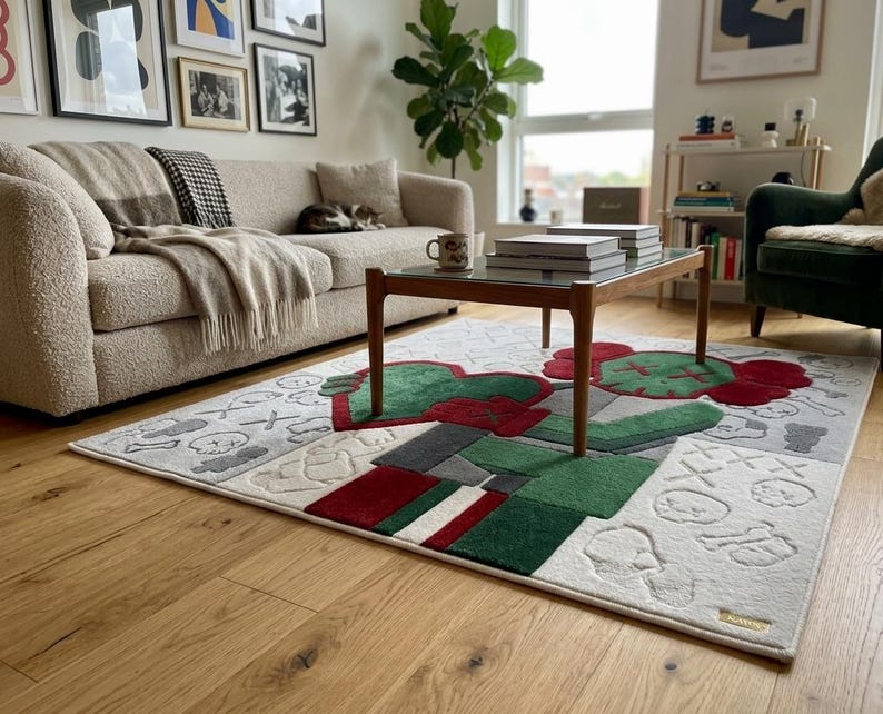 May include: A rectangular area rug featuring a large, stylised character with red and green sections, set against a light grey background with embossed skull and cross-eye motifs. The design includes geometric blocks of red, green, and white.