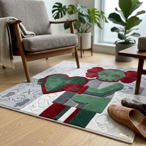 May include: A rectangular area rug featuring a stylised cartoon character in shades of red, green, and grey. The design includes a large red and green figure holding a heart, surrounded by smaller skull and cross-bone motifs on a light grey background.