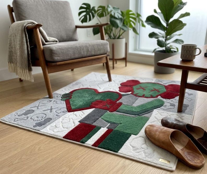 May include: A rectangular area rug featuring a stylised cartoon character in shades of red, green, and grey. The design includes a large red and green figure holding a heart, surrounded by smaller skull and cross-bone motifs on a light grey background.
