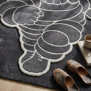 Puede incluir: Una alfombra gris oscuro con un dise&ntilde;o estilizado de una figura con contornos blancos. La alfombra se combina con un par de zapatillas de cuero marr&oacute;n, un libro y una taza. El dise&ntilde;o de la alfombra recuerda a una famosa mascota.