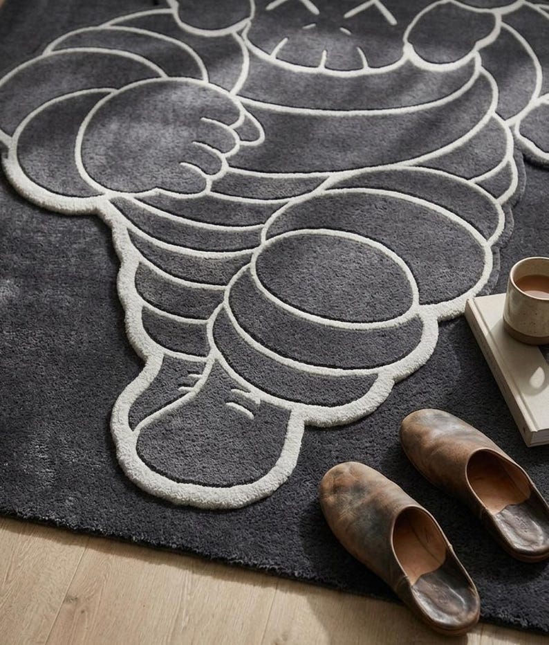 Puede incluir: Una alfombra gris oscuro con un dise&ntilde;o estilizado de una figura con contornos blancos. La alfombra se combina con un par de zapatillas de cuero marr&oacute;n, un libro y una taza. El dise&ntilde;o de la alfombra recuerda a una famosa mascota.
