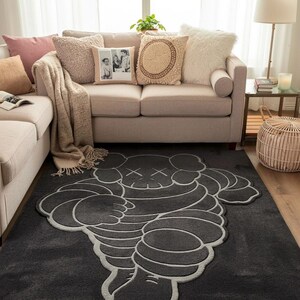 Puede incluir: Una alfombra gris oscuro con un dise&ntilde;o de personaje de dibujos animados estilizado con contornos blancos. La alfombra est&aacute; en un sal&oacute;n con un sof&aacute; beige, cojines y una mesa auxiliar con una l&aacute;mpara. Dimensiones: aprox. 180 cm x 120 cm.