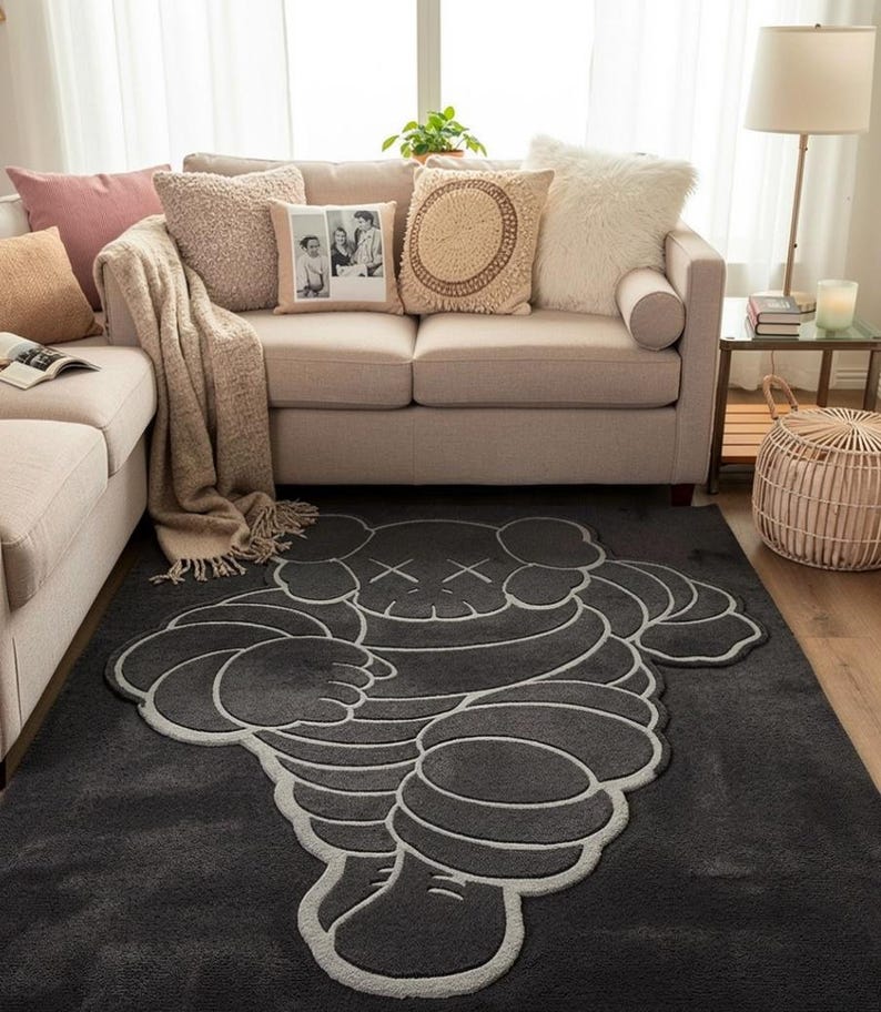 Puede incluir: Una alfombra gris oscuro con un dise&ntilde;o de personaje de dibujos animados estilizado con contornos blancos. La alfombra est&aacute; en un sal&oacute;n con un sof&aacute; beige, cojines y una mesa auxiliar con una l&aacute;mpara. Dimensiones: aprox. 180 cm x 120 cm.