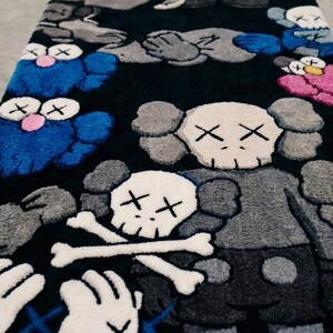 May include: A rectangular rug with a black background featuring a repeating pattern of cartoon-style characters with X-shaped eyes. The design includes blue, grey, pink, and white figures, some with skull-like heads and crossed bones motifs.