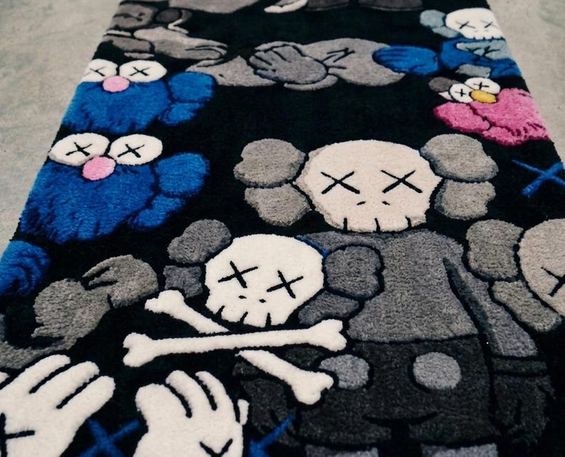 May include: A rectangular rug with a black background featuring a repeating pattern of cartoon-style characters with X-shaped eyes. The design includes blue, grey, pink, and white figures, some with skull-like heads and crossed bones motifs.