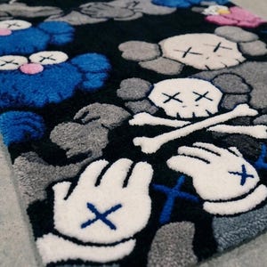 May include: A rectangular tufted rug featuring a black background with a repeating pattern of blue and gray cartoon-style characters. The design includes white gloved hands with blue cross-stitch details and white skull motifs with crossbones.