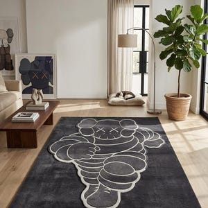 Puede incluir: Una alfombra rectangular gris oscuro con un dise&ntilde;o de figura de dibujos animados estilizada en blanco. La alfombra est&aacute; colocada sobre un suelo de madera en una habitaci&oacute;n luminosa. Obras de arte y una planta en maceta son visibles en el fondo.