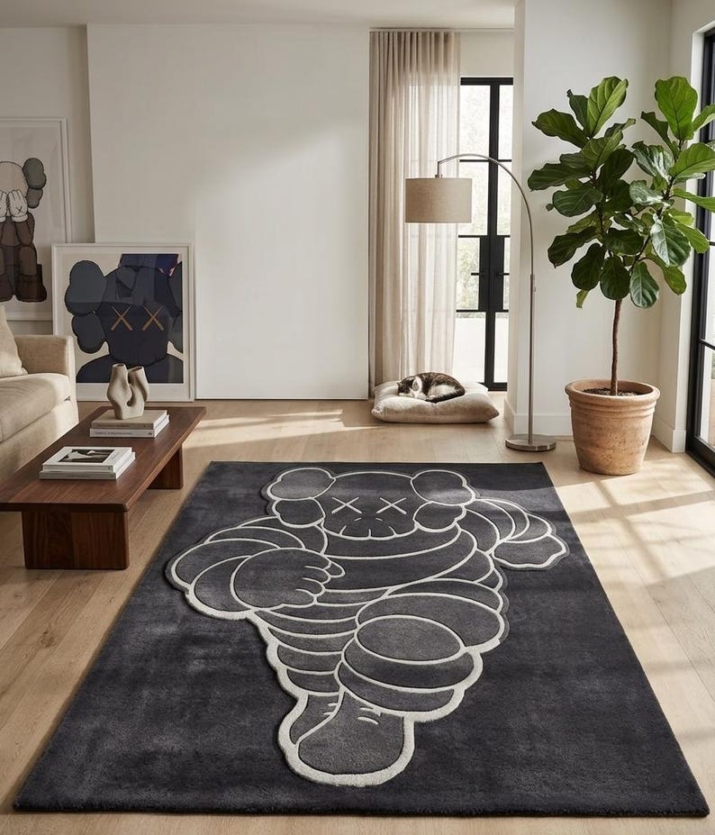 Puede incluir: Una alfombra rectangular gris oscuro con un dise&ntilde;o de figura de dibujos animados estilizada en blanco. La alfombra est&aacute; colocada sobre un suelo de madera en una habitaci&oacute;n luminosa. Obras de arte y una planta en maceta son visibles en el fondo.