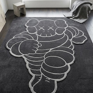 Puede incluir: Una alfombra grande de color gris oscuro con un dise&ntilde;o de personaje de dibujos animados estilizado con contornos blancos. La alfombra tiene una textura afelpada y est&aacute; colocada sobre un suelo gris oscuro. El personaje tiene los ojos tachados.