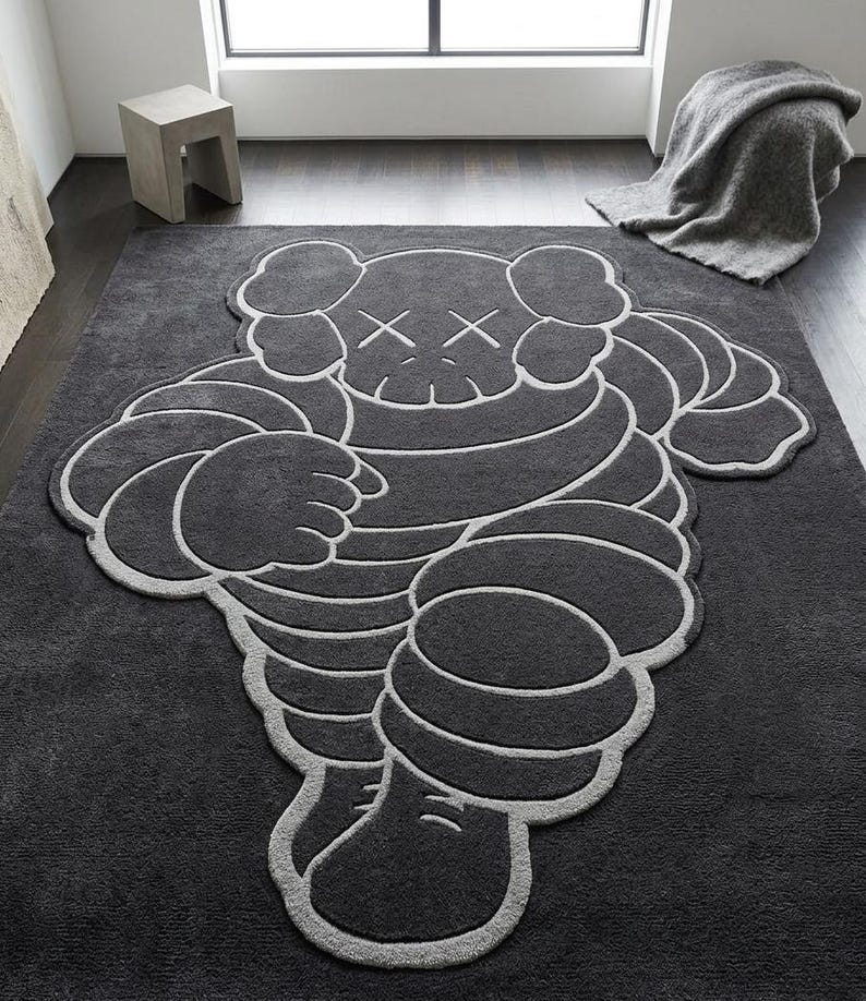 Puede incluir: Una alfombra grande de color gris oscuro con un dise&ntilde;o de personaje de dibujos animados estilizado con contornos blancos. La alfombra tiene una textura afelpada y est&aacute; colocada sobre un suelo gris oscuro. El personaje tiene los ojos tachados.