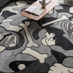 May include: A large area rug featuring a repeating abstract pattern of bones and organic shapes in shades of black, charcoal grey, light grey, and off-white. The design consists of thick, tufted lines that create a textured, layered appearance.