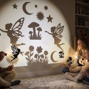 Op de afbeelding: Een set zwarte silhouet schaduwpoppen op stokjes, waaronder feeën, paddenstoelen, bloemen, sterren en manen. De poppen worden voor een lichtbron gehouden om donkere vormen op een muur te projecteren, wat een scène met vliegende feeën creëert.