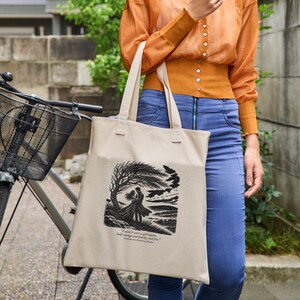 May include: A beige canvas tote bag featuring a black ink-style illustration of two figures embracing under a windswept tree. Below the image, the text reads: I wish I were a girl again, half-savage and hardy, and free. - Emily Bronte, Wuthering Heights.